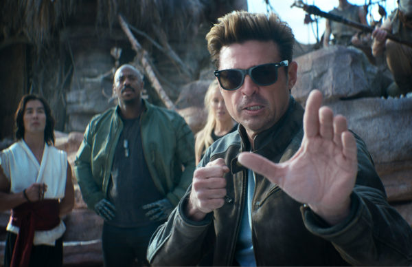 A scene from 'Mortal Kombat 2' featuring a group of diverse characters, with one man in the foreground wearing sunglasses and striking a fighting pose, while others observe in the background.