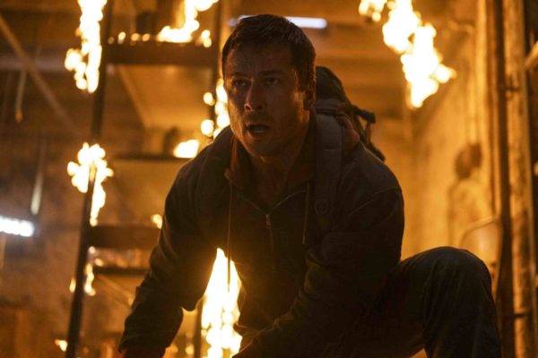 Glen Powell appears as a character in a scene from the Paramount Pictures film "The Running Man," with flames and smoke in the background, conveying a dramatic and intense moment.