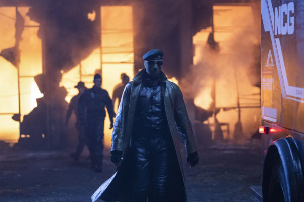 A scene from 'The Running Man' featuring a main character in a long, golden coat and sunglasses, walking confidently towards the camera, with shadows of other figures behind him and an orange glow from a fire illuminating the background.