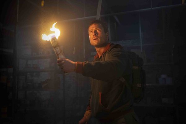 Glen Powell, holding a lit torch, appears determined in a dimly lit environment, suggesting suspense and action from the film 'The Running Man'.