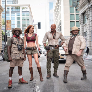 A scene from Jumanji 3 features four characters: Franklin 'Mouse' Finbar, Ruby Roundhouse, Dr. Smolder Bravestone, and Professor Shelly Oberon, standing in a city street, dressed in adventure-themed outfits ready for action.