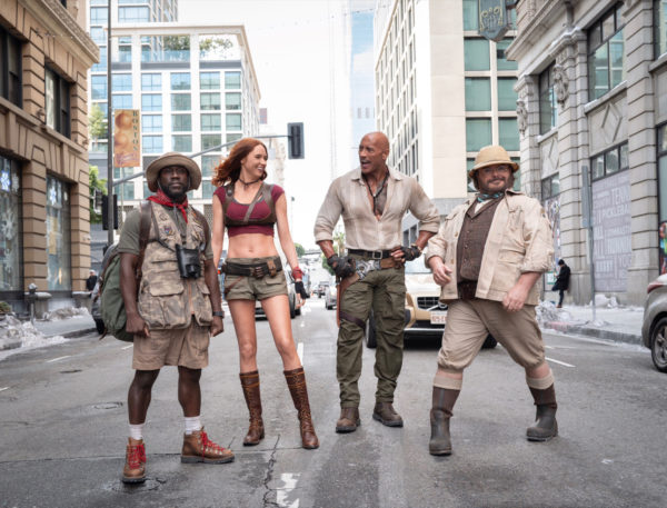 A scene from Jumanji 3 features four characters: Franklin 'Mouse' Finbar, Ruby Roundhouse, Dr. Smolder Bravestone, and Professor Shelly Oberon, standing in a city street, dressed in adventure-themed outfits ready for action.
