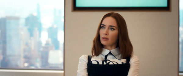 Emily Blunt as Emily Charlton in a scene from 'The Devil Wears Prada 2,' wearing a stylish outfit with a black and white motif, seated in a modern office setting with a city skyline visible through the window.