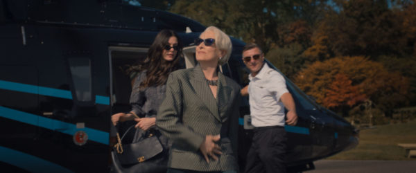 In a scene from 'The Devil Wears Prada 2', Meryl Streep as Miranda Priestly exits a helicopter, dressed in a stylish green striped suit and sunglasses, flanked by Anne Hathaway as Andie Sachs and a crew member. The backdrop features autumn trees in vibrant colors.