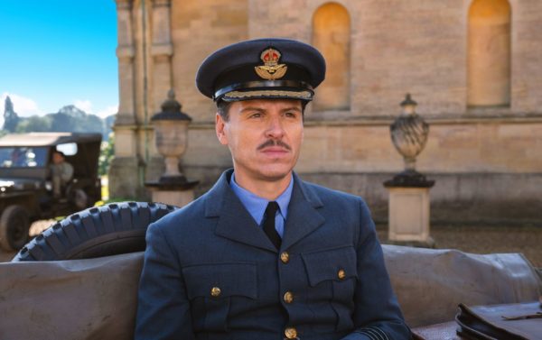 Andrew Scott as Captain James Stagg in the film 'PRESSURE,' wearing a military uniform and cap, seated in front of a historic building with a truck visible in the background.