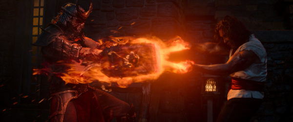 A dramatic scene from Mortal Kombat II, featuring two characters in combat. One character, wearing a fearsome armor with a helmet and holding a flaming weapon, displays intense energy as it clashes with the other character, who is casting a fiery spell. The background includes a dimly lit stone structure.
