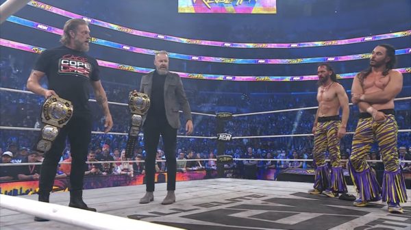 Adam Copeland and Christian Cage stand in a wrestling ring, holding the AEW Tag Team Titles, staring down the Young Bucks. The atmosphere is electric, with a large crowd in the background.