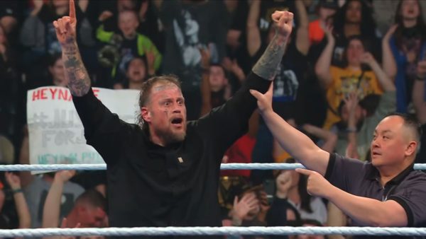 A victorious wrestler raises his arm in celebration after winning a match at WWE SmackDown, with the referee beside him signaling the win. The crowd in the background is cheering enthusiastically.