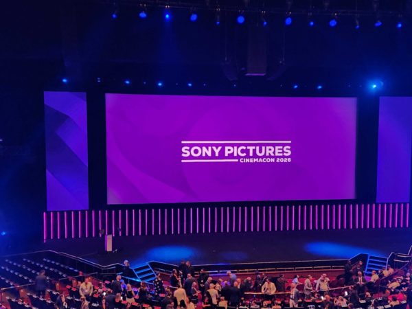 A stage set for a Sony Pictures presentation at CinemaCon 2026, featuring a large purple backdrop with the Sony Pictures logo and a podium on stage. The audience is seated and chatting in the auditorium, with dynamic lighting creating an energetic atmosphere.