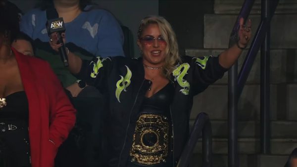 The AEW World Champion Thekla stands confidently with her arms raised and a championship belt around her waist, wearing sunglasses and a black jacket adorned with green flames, taunting her challenger Alex Windsor during a wrestling event.