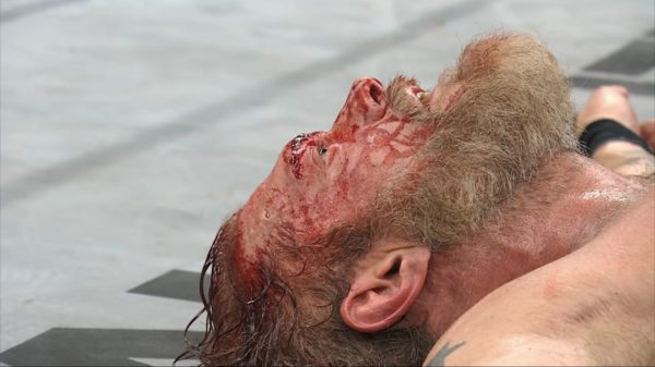 A bloodied Adam Copeland lies on the ground, appearing defeated after a match against FTR at AEW Dynasty. His expression reflects exhaustion and defeat, with blood visibly covering his face and chest.