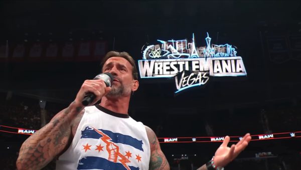 CM Punk passionately delivers a promo while standing on an announce table, with a large WrestleMania sign illuminated in the background. He is wearing a white tank top featuring a blue and red design, showcasing his tattoos.