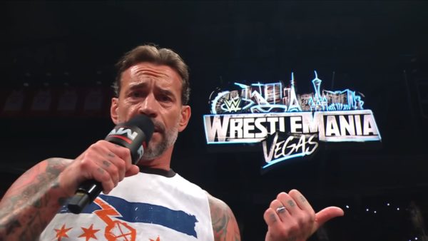 A professional wrestler passionately addresses the crowd while holding a microphone, with the WrestleMania Las Vegas logo prominently displayed in the background.