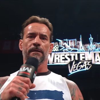 CM Punk stands on stage speaking into a microphone, expressing his views while a large WrestleMania logo is illuminated behind him.