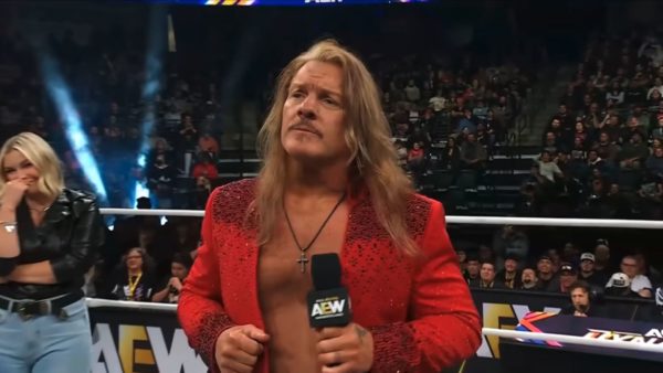 Chris Jericho, wearing a sparkly red jacket, stands in the wrestling ring at AEW Dynamite. He holds a microphone and appears to be confronting an unseen opponent, with a female figure laughing in the background.