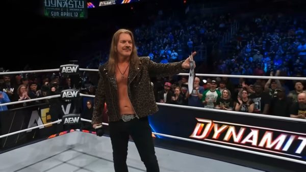 Chris Jericho stands in a wrestling ring, wearing a leopard-print jacket, holding a contract and posing for the crowd at AEW Dynamite. The background features excited fans and an arena filled with lights.