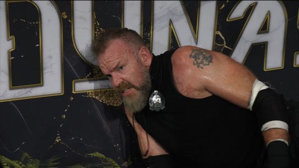 A distressed wrestler, Christian Cage, appears exhausted and disheveled after competing at a wrestling event, with a backdrop featuring the word "Dynasty." He has a noticeable beard and tattoos, and sweat glistens on his skin, indicating an intense match.