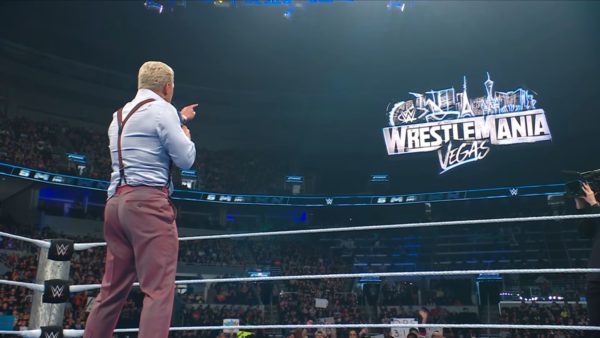 A wrestler delivers a passionate promo on stage, pointing toward a large digital display featuring the "WrestleMania Vegas" logo in the background, with an enthusiastic crowd visible in the arena.