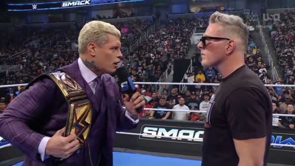 Cody Rhodes, dressed in a purple suit and holding a championship belt, confronts Pat McAfee, who is wearing a black t-shirt and glasses. Both are engaged in an intense discussion in the ring during WWE SmackDown, with a large audience in the background.
