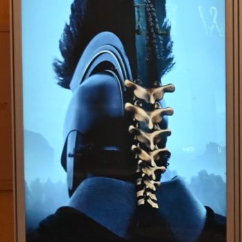 A promotional poster for the film 'The Odyssey,' featuring a close-up of a warrior's helmet with a striking mohawk design, set against a blue background. The poster includes a release date of July 17, 2026, and is directed by Christopher Nolan.