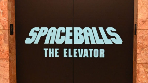 A black elevator door features bold, light blue text reading 'SPACEBALLS THE ELEVATOR'.