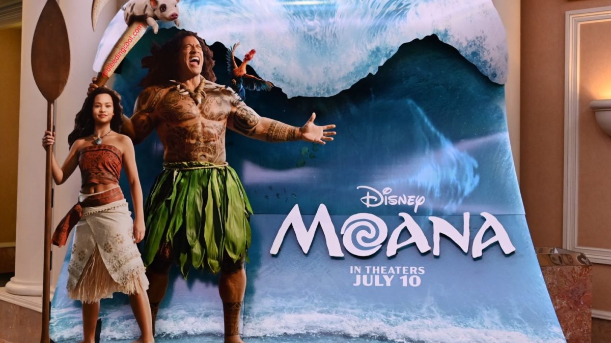 A promotional display for Disney's 'Moana' features the characters Moana and Maui against a backdrop of a large wave. Moana stands with a spear, while Maui is depicted with tattoos and a large hook, showcasing vibrant colors and a tropical theme.