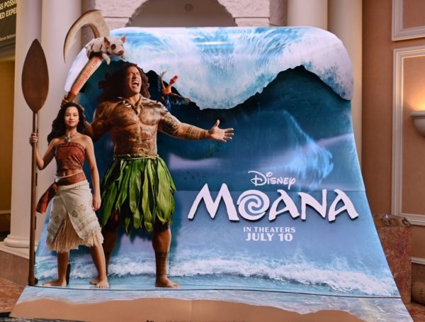 A promotional display for Disney's 'Moana' features the characters Moana and Maui against a backdrop of a large wave. Moana stands with a spear, while Maui is depicted with tattoos and a large hook, showcasing vibrant colors and a tropical theme.