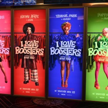 NEON - A promotional display for the film 'I Love Boosters' featuring four characters, each in vibrant costumes against colorful backgrounds—pink, red, blue, and green. The characters are portrayed by Keke Palmer, Naomi Ackie, Taylour Paige, and Poppy Liu, with text announcing the film's release on May 22.
