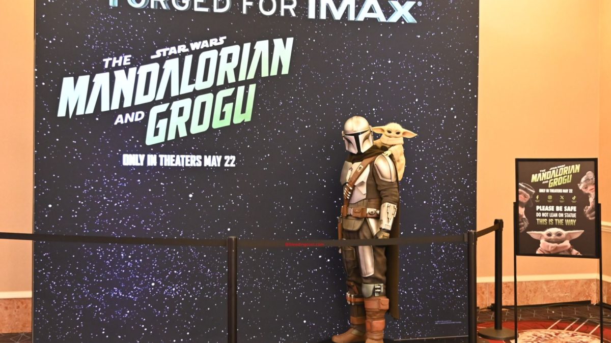 A life-sized standee of the Mandalorian, with Grogu on his back, is set against a starry backdrop promoting "The Mandalorian and Grogu," with a sign reminding guests to not lean on the statue.