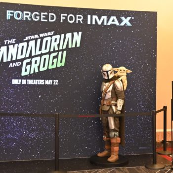 A life-sized standee of the Mandalorian, with Grogu on his back, is set against a starry backdrop promoting "The Mandalorian and Grogu," with a sign reminding guests to not lean on the statue.