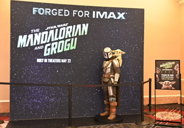 A life-sized standee of the Mandalorian, with Grogu on his back, is set against a starry backdrop promoting "The Mandalorian and Grogu," with a sign reminding guests to not lean on the statue.