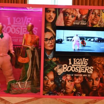 NEON - A promotional display for the film 'I Love Boosters,' featuring two mannequins dressed in colorful outfits, including a pink oversized hoodie. The display includes a screen showing a scene from the movie and artwork featuring various characters with vibrant hairstyles and makeup, set against a pink background.