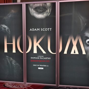 A promotional display for the film 'Hokum,' featuring the names of cast member Adam Scott and director Damian McCarthy. The design includes dark, eerie graphics that hint at horror elements, with a focus on unsettling facial expressions.