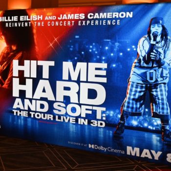 A promotional display for the concert experience 'Hit Me Hard and Soft' featuring Billie Eilish and James Cameron, with vibrant graphics and the event date May 8 prominently featured.