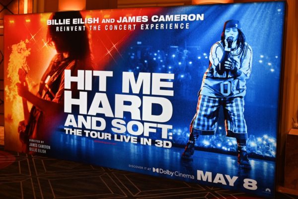 A promotional display for the concert experience 'Hit Me Hard and Soft' featuring Billie Eilish and James Cameron, with vibrant graphics and the event date May 8 prominently featured.