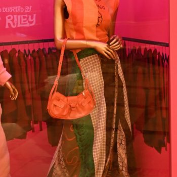 NEON - A stylish mannequin dressed in a colorful outfit featuring a bright striped top and a flowing skirt with various patterns. The display includes an orange handbag and a floral-patterned bag at the base, all set against a vibrant pink backdrop emphasizing a fashion theme.