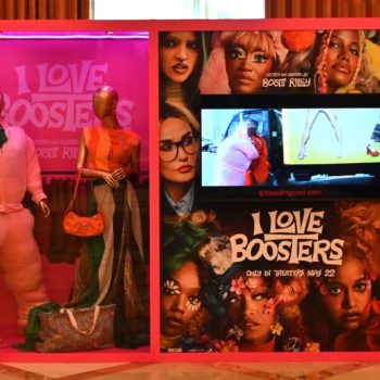 NEON - A display for the movie 'I Love Boosters' featuring two mannequins dressed in vibrant, whimsical outfits alongside a monitor showing scenes from the film. The background is prominently adorned with colorful promotional imagery and the movie title, all presented in a pink-themed setting.