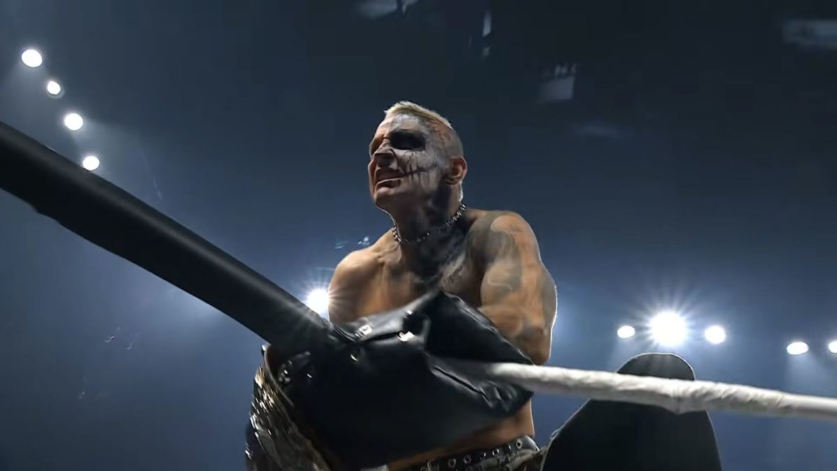 Darby Allin, a professional wrestler with distinctive face paint and tattoos, celebrates with the AEW Championship belt in hand, standing near the ring ropes under bright arena lights.