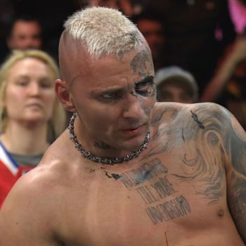 Darby Allin, a professional wrestler with distinctive tattoos and a mohawk hairstyle, appears exhausted after a tough win against Andrade El Idolo. Blood is visible on his lip, and fans can be seen cheering in the background.