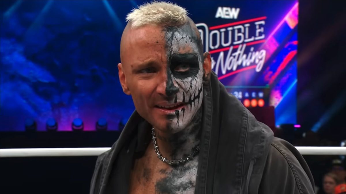 A wrestler with a half-painted face, showcasing a skull design, expresses intensity and determination in the wrestling ring, wearing a dark jacket and a chain necklace, with a dynamic event backdrop behind him.