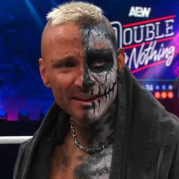 A wrestler with a half-painted face, showcasing a skull design, expresses intensity and determination in the wrestling ring, wearing a dark jacket and a chain necklace, with a dynamic event backdrop behind him.