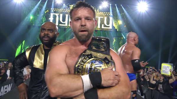 A wrestler, shirtless and carrying a championship belt, stands prominently in the foreground with a look of determination. Another man in a black and gold jacket appears surprised in the background, while a third wrestler is partially visible. Fans are cheering with signs in a vibrant arena setting.