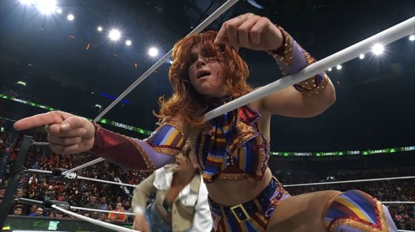 A female wrestler, Jamie Hayter, appears upset after an event at AEW Dynasty, pointing with determination while displaying a vibrant, multicolored outfit adorned with studs. The crowd is visible in the background, adding to the dramatic atmosphere.