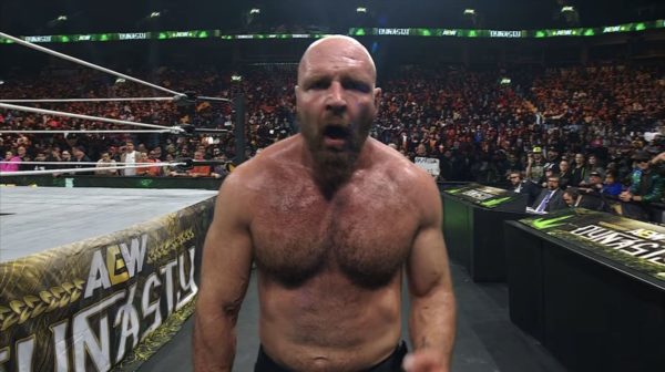 A muscular bald man, Jon Moxley, stands in the ring following a match, visibly emotional and expressing intensity. The audience is filled with fans cheering and holding signs in the background.