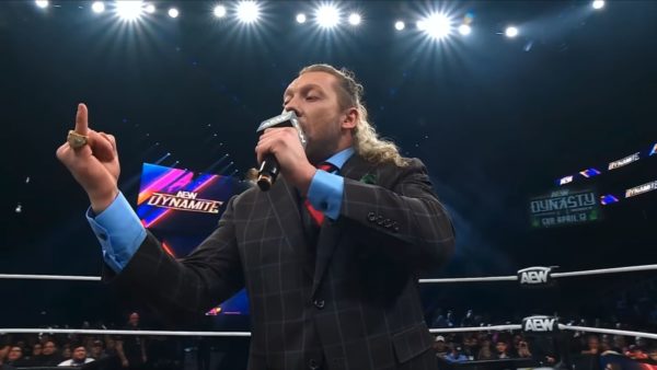 Kenny Omega holds a microphone and shows off MJF's Dynamite Diamond Ring while addressing the audience at AEW Dynamite, with bright lights shining above and fans in the background.