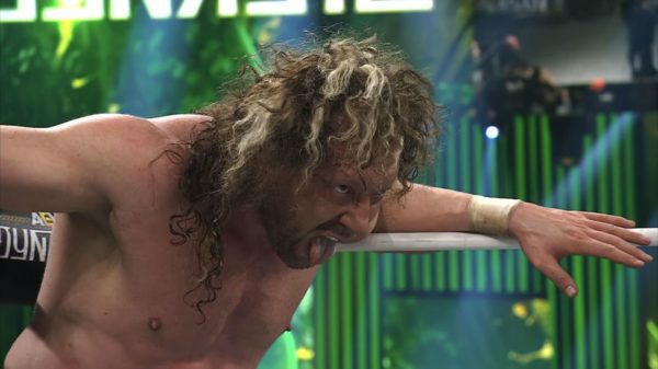 Kenny Omega, visibly intense with a fierce expression, hangs onto the ropes of the wrestling ring, preparing for his title challenge at AEW Dynasty. He has long, curly hair and is shirtless, showing the physical strain of the match.