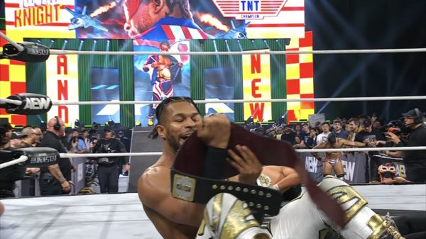 A triumphant wrestler, Kevin Knight, celebrates while holding the TNT Championship belt in a wrestling ring, surrounded by fans and a vibrant backdrop featuring the words 'AND NEW' alongside an impressive graphic.