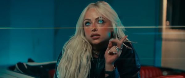 A young woman with long blonde hair gestures with her hand while making a thoughtful expression, set against a blue background. She wears a black leather jacket and has multiple rings on her fingers.