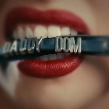 Close-up of a woman's mouth with bold red lipstick biting down on a choker that reads 'Daddy Dom'. The background is blurred, emphasizing the focus on the choker and lips.