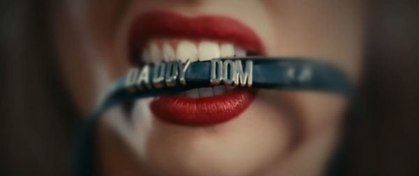 Close-up of a woman's mouth with bold red lipstick biting down on a choker that reads 'Daddy Dom'. The background is blurred, emphasizing the focus on the choker and lips.